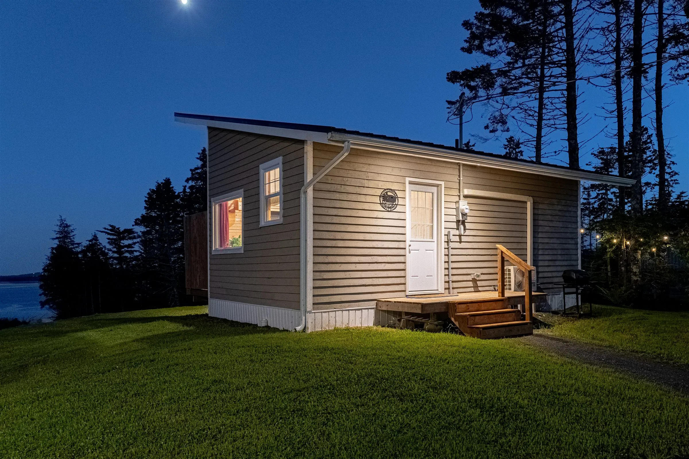 The cabin at twilight under the moon, with lit entry steps and the inlet beyond the trees.