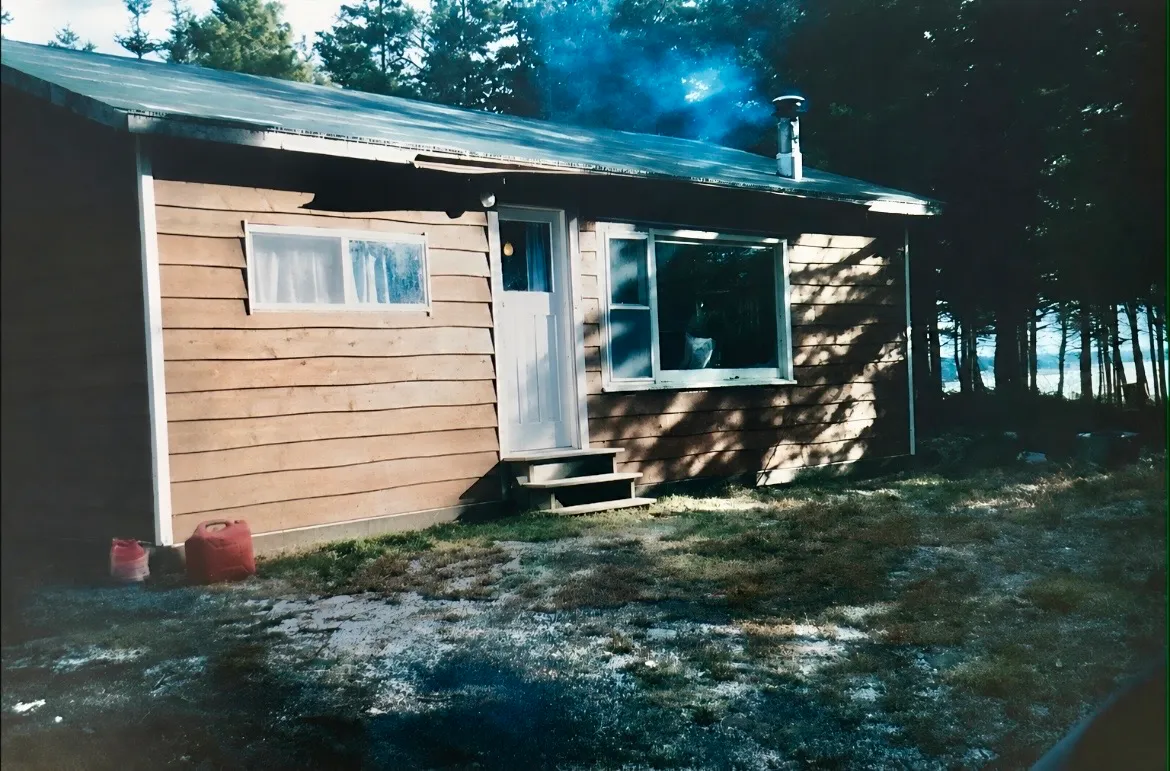 The original cabin before renovations - rough board siding, a wood-stove chimney, and the inlet through the trees.