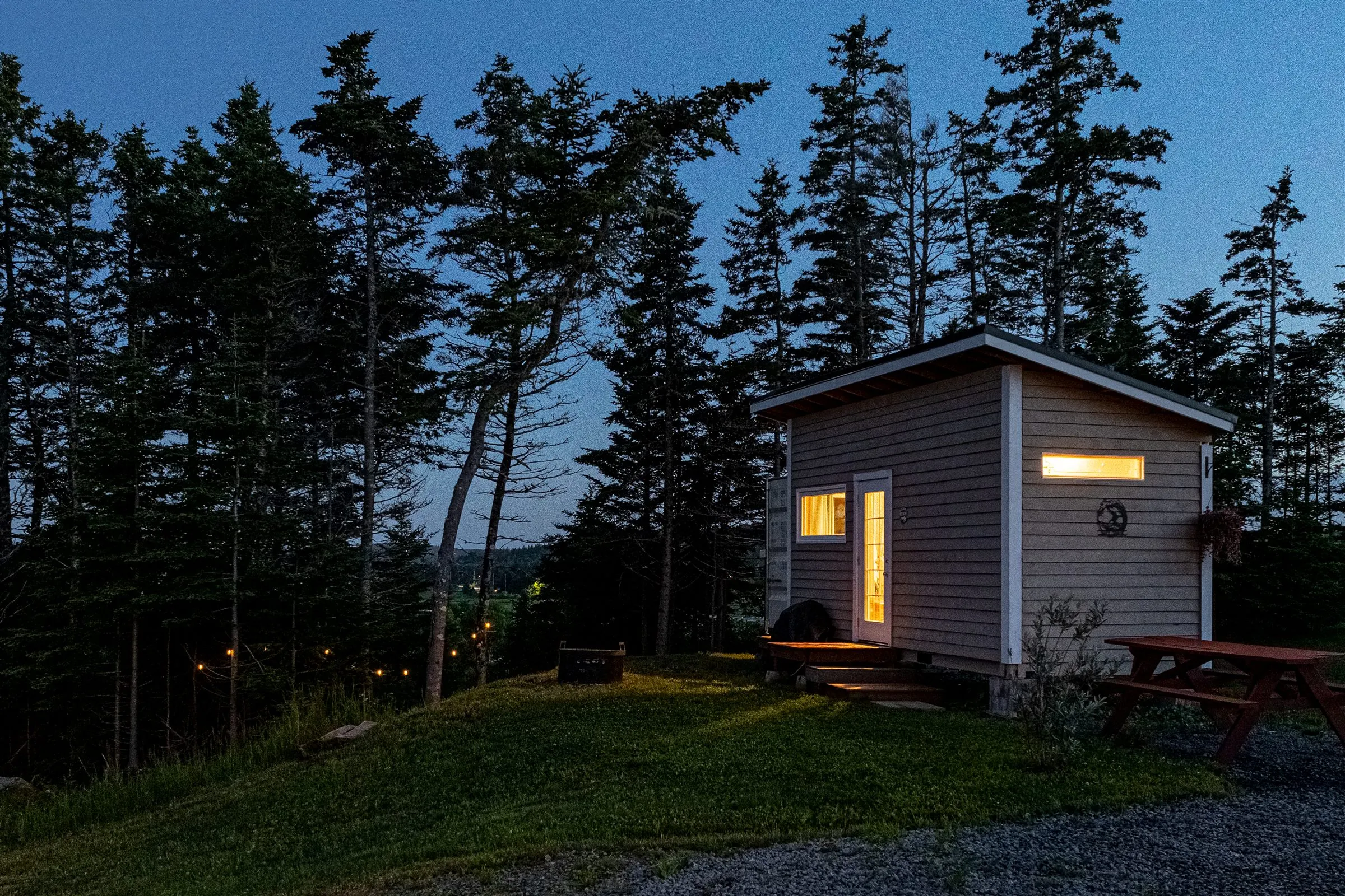 The Surf Shack bunkie glowing warmly at dusk, with a picnic table beside it and string lights trailing down the hill.