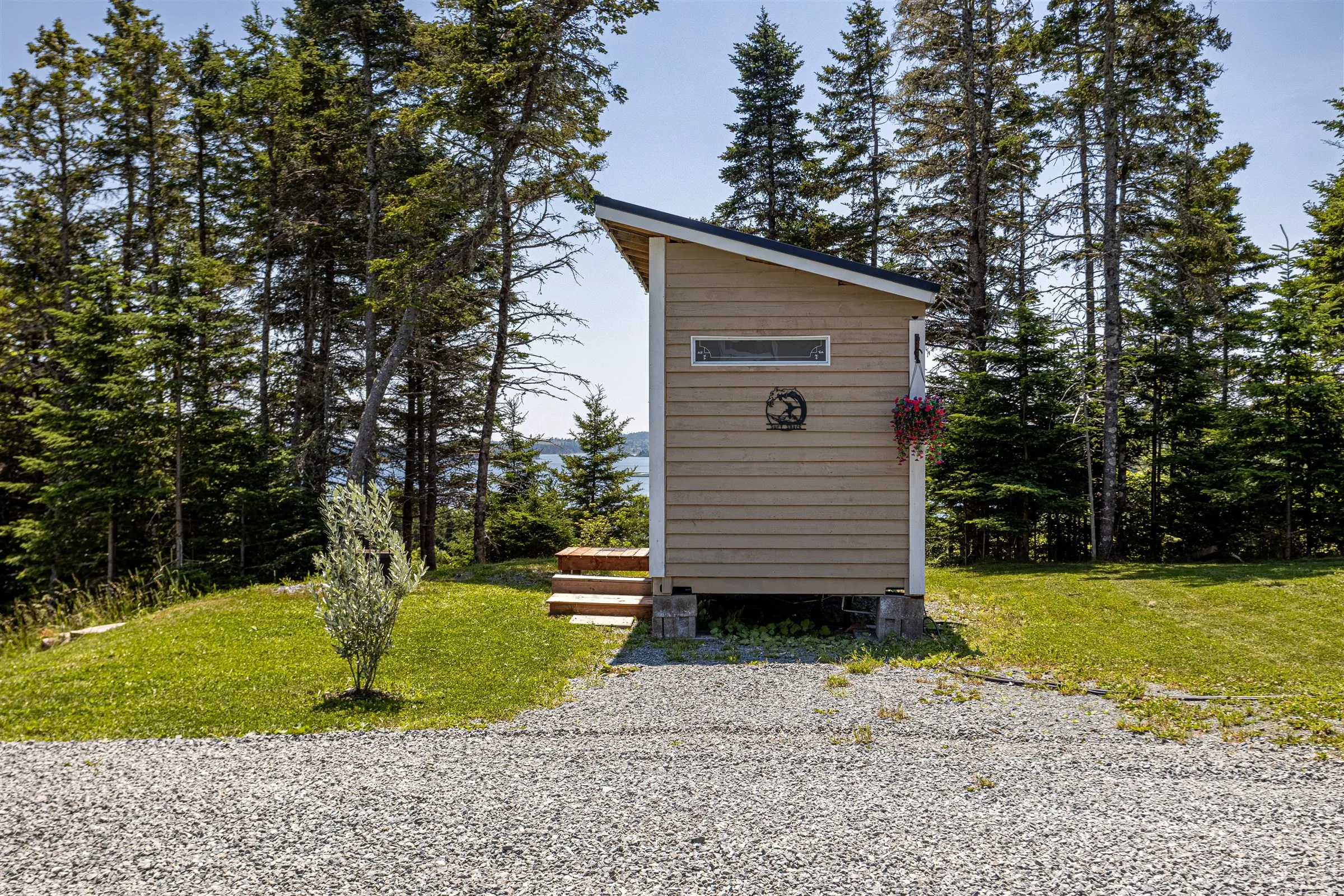 The bunkie's end wall with a hanging flower basket, seen from the gravel driveway.