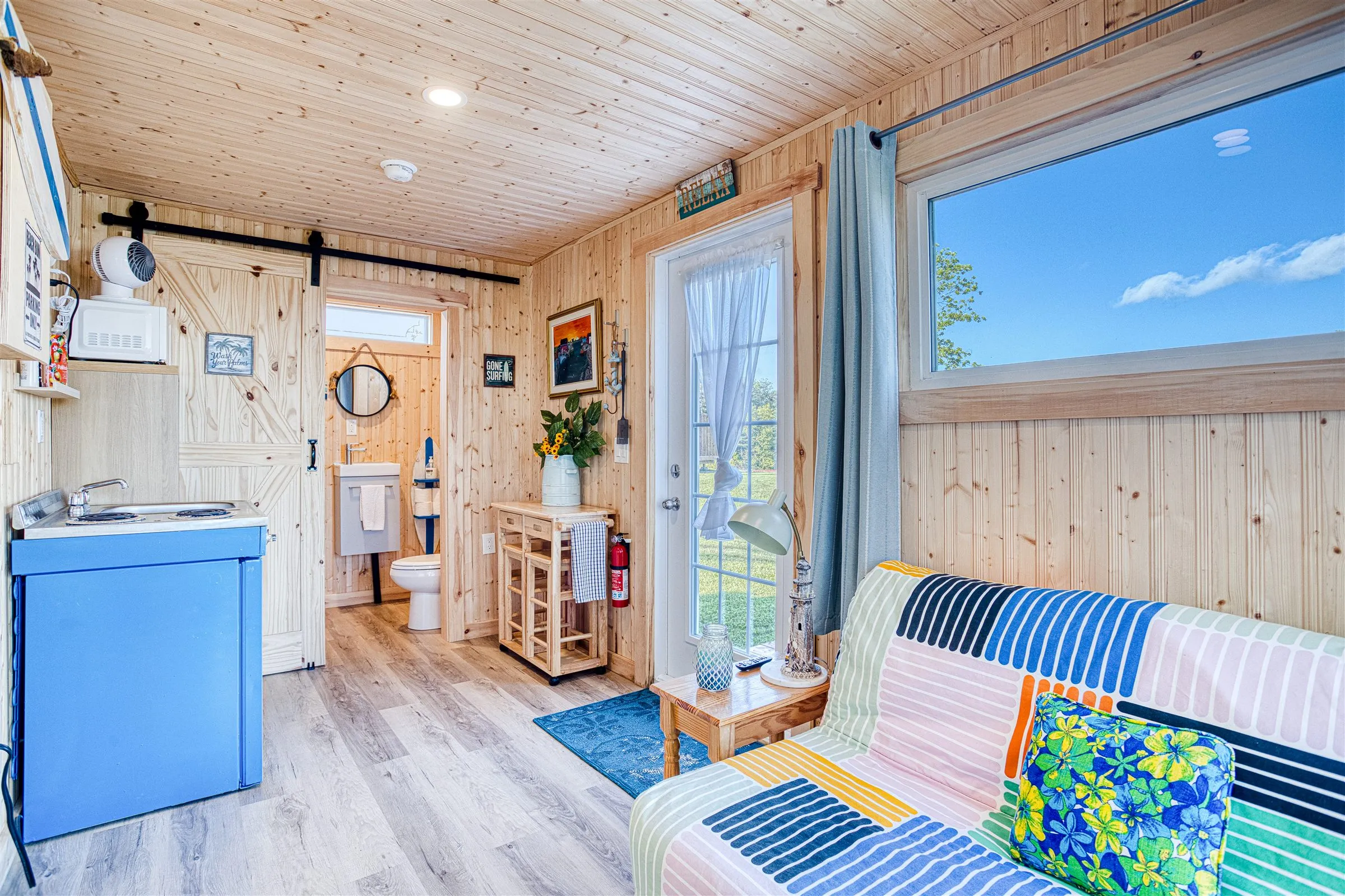 Open-plan interior showing the striped futon, blue kitchenette, and barn door open to the bathroom.